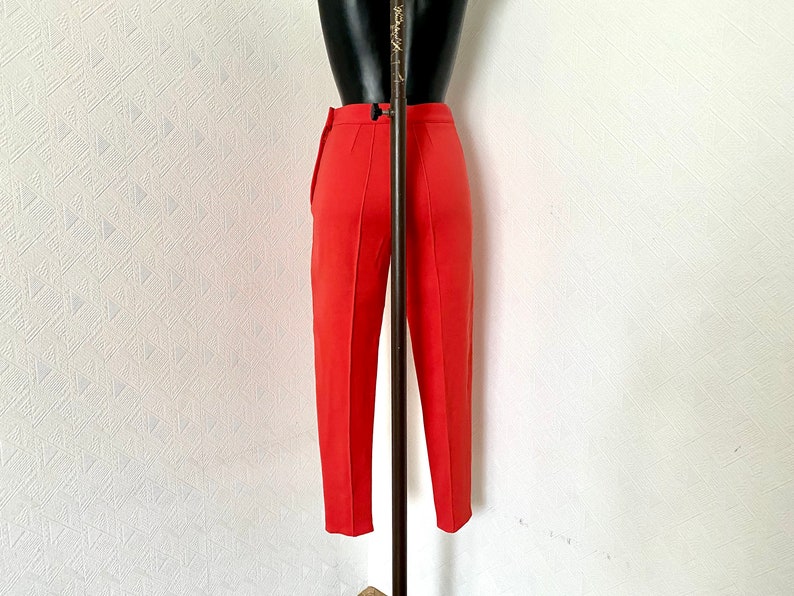 May include: A pair of red, straight-leg pants with a center crease. The pants are on a mannequin and are hanging on a white wall.