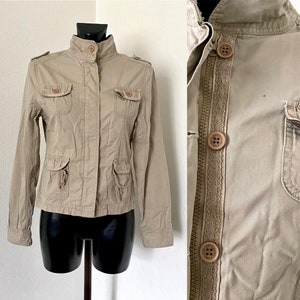 Può includere: Giacca beige con collo alto, quattro tasche frontali e chiusura con bottoni. La giacca ha maniche lunghe e spalline. La giacca è esposta su un manichino nero.