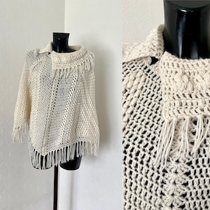 May include: A white crocheted poncho with a fringe border. The poncho has a high neck and a loose fit.