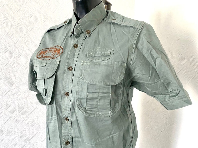 Pu&ograve; includere: Una camicia verde a maniche corte con bottoni, pi&ugrave; tasche e una toppa ricamata marrone che dice "African Wildlife Safaris" sul petto sinistro.