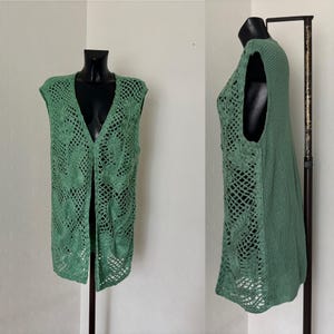 May include: A sage green, open-front vest with a V-neck. The vest features a crocheted design with floral and geometric patterns. The sleeveless garment is displayed on a black mannequin, showcasing the intricate openwork and texture of the knit.