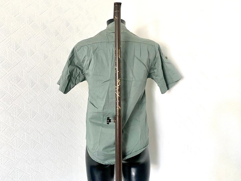 Pu&ograve; includere: Una camicia verde salvia a maniche corte con bottoni su un manichino.