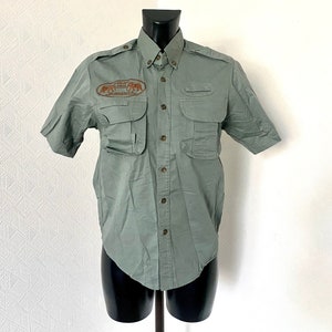 Pu&ograve; includere: Una camicia verde a maniche corte con bottoni e una toppa sul petto sinistro che dice "African Wildlife Safaris" e raffigura un'immagine di elefanti. La camicia ha diverse tasche.