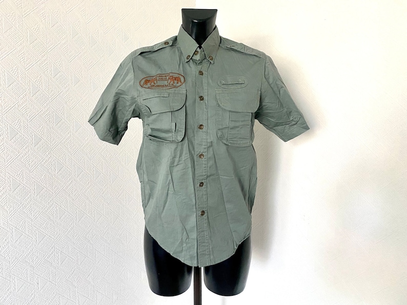 Pu&ograve; includere: Una camicia verde a maniche corte con bottoni e una toppa sul petto sinistro che dice "African Wildlife Safaris" e raffigura un'immagine di elefanti. La camicia ha diverse tasche.