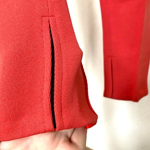 May include: Close-up of a pair of coral red pants with black trim along the seams. The pants have a slit at the bottom of each leg.