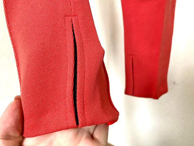 May include: Close-up of a pair of coral red pants with black trim along the seams. The pants have a slit at the bottom of each leg.