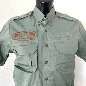 Pu&ograve; includere: Una camicia verde a maniche corte con bottoni e diverse tasche. La camicia ha una toppa ricamata sul petto sinistro che recita "Elephant Hills Thailand - Safari around the World - Elephant Hills Club".