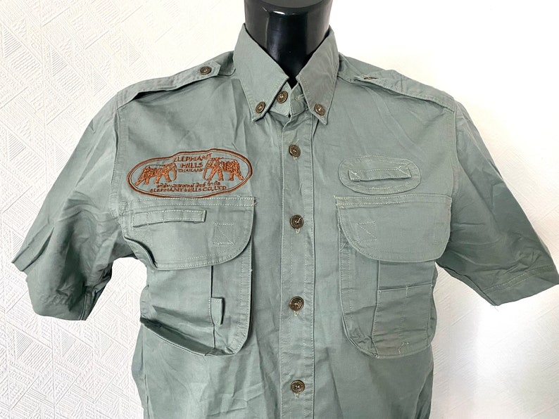 Pu&ograve; includere: Una camicia verde a maniche corte con bottoni e diverse tasche. La camicia ha una toppa ricamata sul petto sinistro che recita "Elephant Hills Thailand - Safari around the World - Elephant Hills Club".