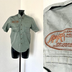 Pu&ograve; includere: Una camicia verde a maniche corte con bottoni, due tasche a toppa e un logo ricamato marrone che dice "Elephant Hills Thailand" sul petto sinistro.