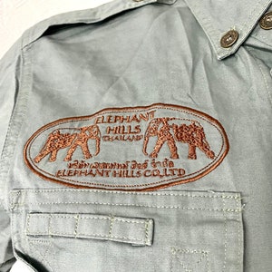 Pu&ograve; includere: Una camicia a maniche lunghe verde chiaro con un logo ricamato marrone che dice "Elephant Hills Thailand" e presenta due elefanti. Il logo include anche il testo "Elephant Hills Co. Ltd."