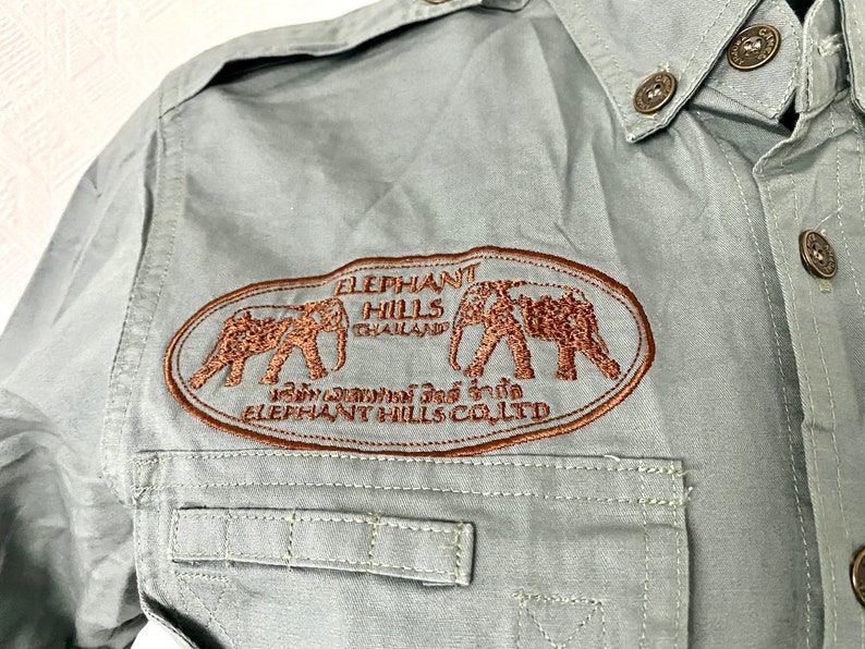 Pu&ograve; includere: Una camicia a maniche lunghe verde chiaro con un logo ricamato marrone che dice "Elephant Hills Thailand" e presenta due elefanti. Il logo include anche il testo "Elephant Hills Co. Ltd."