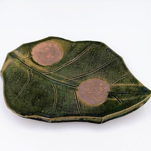 May include: A green ceramic leaf-shaped dish with two raised brown circles. The dish has a textured surface and a slightly irregular edge.