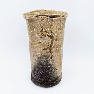 May include: A tall, cylindrical ceramic vase with a textured, off-white glaze. The vase has a slightly irregular shape and a dark brown base.