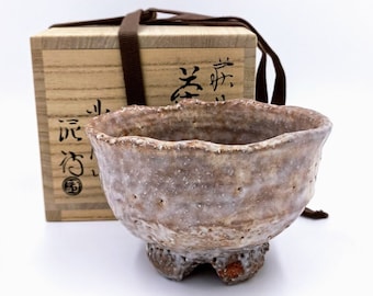 May include: A small, rustic, brown and white ceramic teacup with a rough, textured surface. The cup has three small feet and is sitting on a wooden box with Japanese characters.