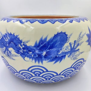 Puede incluir: Un gran cuenco de cerámica blanca con un diseño de dragón azul y blanco. El cuenco tiene un borde festoneado y un patrón de ondas azul y blanco alrededor de la base.