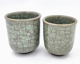 May include: Two green ceramic teacups with a crackle glaze. The cups have a light green base and a darker green crackle glaze.