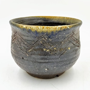 May include: A small, dark gray ceramic pot with a brown glaze and a geometric pattern. The pot has a slightly rounded shape and a wide opening.