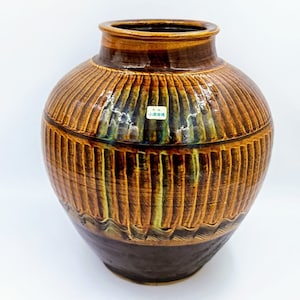 Puede incluir: Un jarrón grande de cerámica marrón con una superficie texturizada y nervada. El jarrón tiene un esmalte marrón oscuro con toques de verde y amarillo. El jarrón mide aproximadamente 30 cm de alto.