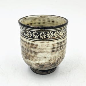 May include: A white ceramic cup with a black and white floral design around the rim. The cup has a textured, slightly rough surface.
