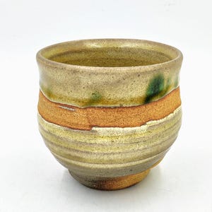 May include: A small, hand-thrown ceramic cup with a light brown glaze and a band of orange glaze around the middle. The cup has a slightly rounded shape and a textured surface.