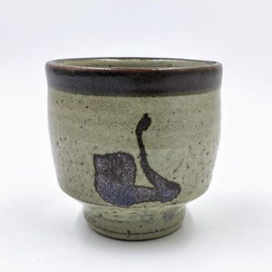 May include: A ceramic cup with a speckled, light green exterior and a dark brown rim. The cup features a dark blue and black abstract design on the side. The cup has a pedestal base.