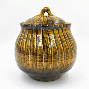 Puede incluir: Un tarro de cerámica con tapa, con un esmalte rayado marrón y amarillo. El tarro tiene un cuerpo redondeado y una abertura ligeramente acampanada.