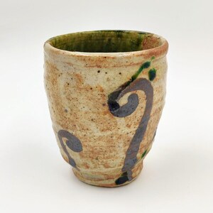 May include: Handmade ceramic cup with a textured, speckled beige exterior. The rim is a darker brown with a green interior. Dark blue and green swirl designs decorate the cup's exterior. A unique piece for beverages or display.