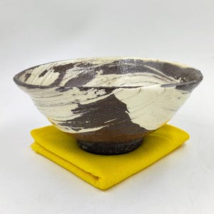 Op de afbeelding: Een witte keramische kom met bruine, draaiende patronen. De kom staat op een gele doek.