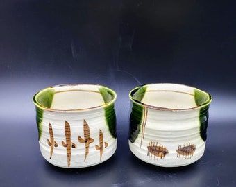 May include: Two ceramic teacups with green and brown glaze. The cups have a white interior and brown painted designs.