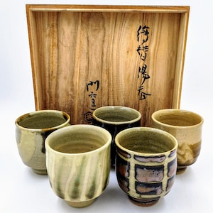 May include: Five ceramic teacups with different glaze patterns, including green, brown, and black, are displayed in a wooden box with Japanese characters. The box is open and the cups are arranged in a semi-circle.