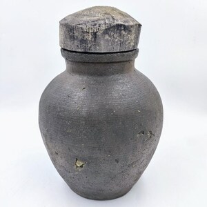 May include: A dark gray ceramic jar with a wooden lid. The jar has a rough, textured surface and appears to be handmade.