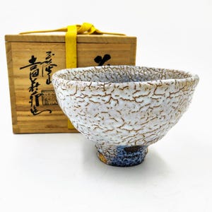 May include: A white ceramic tea bowl with a crackled glaze and a blue and gold base. The bowl is in a wooden box with Japanese characters on the side.
