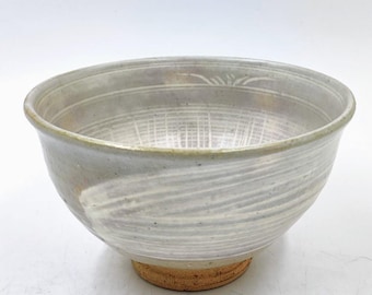 May include: A handmade ceramic bowl with a light gray glaze and a textured, organic design. The bowl features a wide, shallow shape with a slightly flared rim and a textured base. The interior has a grid-like pattern.