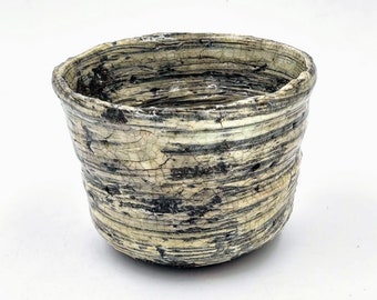May include: A small, white ceramic bowl with a textured, crackled glaze. The bowl has a wide, round opening and a slightly flared rim. The glaze is a light cream color with dark gray streaks and lines.