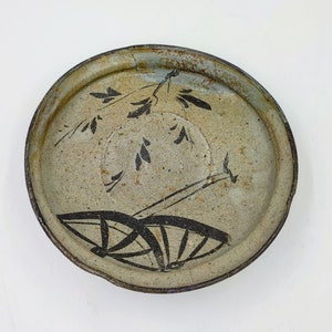 May include: A round ceramic plate with a light gray glaze and black painted designs. The plate features a stylized design of leaves and a fan.