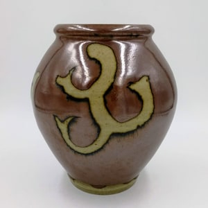 May include: A brown ceramic vase with a stylized design in a light brown color. The vase has a rounded body and a slightly flared opening.