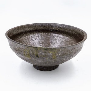 May include: A dark brown ceramic bowl with a textured surface and a flared rim. The bowl is set on a short, rounded base. The interior has a speckled appearance with hints of green. A rustic piece.