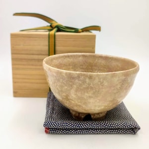 以下が含まれることがあります： 表面がざらざらした薄い茶色の陶器の茶碗。丸みを帯びた形をしていて、青と白の模様の布の上に置かれている。茶碗は緑と金の色のリボンが付いた木箱に入っている。