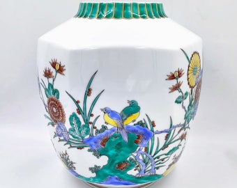May include: A white ceramic vase with a green and gold rim. The vase is decorated with a floral pattern and two birds in blue, yellow, and green.