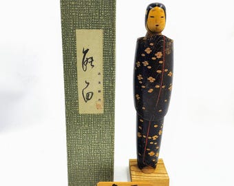 May include: A wooden kokeshi doll with a black kimono featuring a gold floral pattern, standing on a light wood base. Beside it is a tall, textured green storage box with a label containing Japanese text and a small wooden sign with Japanese text.