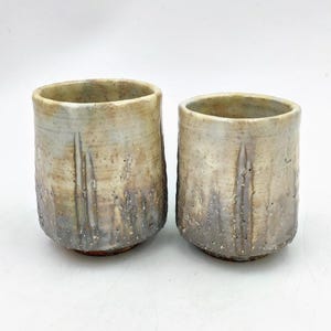 May include: Two ceramic cups with a light brown and gray glaze. The cups have a textured surface and a slightly rounded shape.