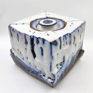 May include: Square ceramic container with a lid. The container is white with blue and brown accents. The lid has a circular design. The ceramic piece has a textured surface and a unique, handcrafted appearance.