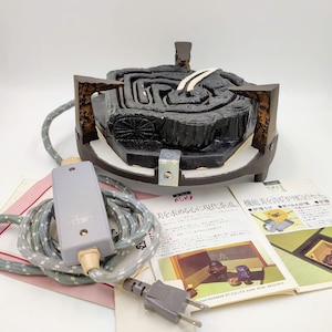 Könnte beinhalten: Ein schwarzer und brauner elektrischer Hibachi-Grill mit Kabel und Stecker. Der Grill steht auf einer weißen Oberfläche. Der Text auf der Bedienungsanleitung lautet "にしき 美を求める心に現代茶道。機能美を誇る「やりセッ"