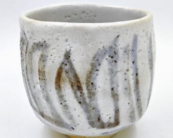 May include: A white ceramic teacup with a brown and grey abstract design. The cup has a rounded shape and a slightly flared rim.