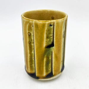 May include: A yellow ceramic cup with vertical lines and a dark green glaze. The cup is handmade and has a rustic look.