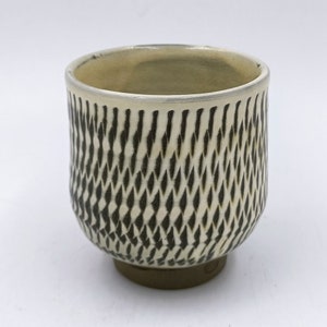 Puede incluir: Una taza de cerámica blanca con un patrón geométrico en blanco y negro. La taza tiene un fondo redondeado y un borde ligeramente acampanado.