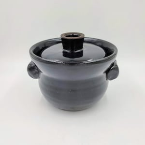 Puede incluir: Una olla de cerámica negra con tapa y dos asas. La olla tiene un esmalte oscuro y una forma ligeramente redondeada.