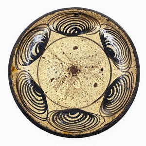 Può includere: Un piatto rotondo in ceramica antica con un centro color crema e un bordo marrone scuro. Il piatto presenta sei disegni a spirale concentrici neri sul bordo. Il centro ha un aspetto macchiato e invecchiato.
