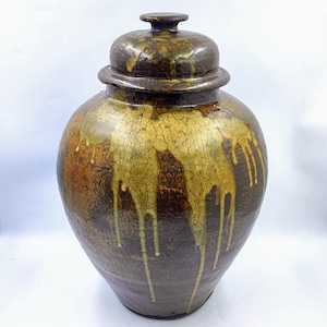Puede incluir: Un jarrón grande de cerámica con tapa. El jarrón es de color marrón oscuro con vetas doradas y anaranjadas. La tapa es de un tono marrón más oscuro. El jarrón tiene una superficie texturizada con efecto craquelado. Es un objeto decorativo.