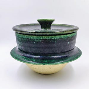 May include: A handcrafted ceramic lidded bowl with a dark green and blue glaze. The bowl has a wide, flat base and a matching lid with a small handle. The lower portion of the bowl is a light beige color. This unique piece is perfect for serving or display.